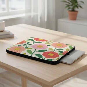 May include: A floral laptop sleeve featuring a cream background with red, pink, and yellow flowers and green leaves. The sleeve has a black border and a laptop is partially inserted. The sleeve is on a light wood table.