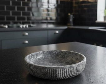 May include: A grey marble bowl with vertical ribbed detailing on the exterior. The bowl sits on a dark countertop in a kitchen setting. The bowl's interior has a marbled pattern.