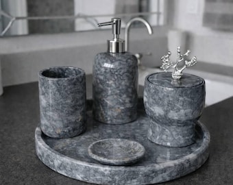 Può includere: Un set da bagno in marmo grigio composto da un dosatore di sapone, un bicchiere, un barattolo con coperchio, un piattino e un vassoio rotondo. Il dosatore di sapone ha una pompa argentata. Il barattolo ha una maniglia decorativa argentata a forma di albero.