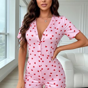 Puede incluir: Mono rosa con estampado de corazones rojos. El mono de manga corta presenta un escote en V y una botonadura frontal. Los bordes de las mangas y los pantalones cortos tienen un ribete festoneado. La prenda est&aacute; hecha de una tela suave y ligera.