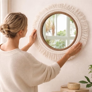 Pode incluir: Espelho redondo com design macramé. Apresenta uma borda de corda natural e franjas creme. O espelho é exibido em uma parede branca. O estilo é boêmio e decorativo.