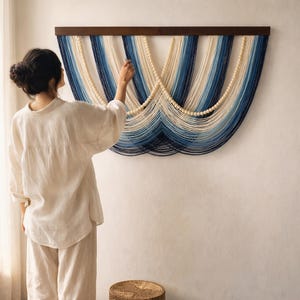 May include: A decorative wall hanging featuring a wooden top bar and a cascade of blue, white, and cream-coloured yarn. The yarn is arranged in vertical stripes and curves, with wooden beads adding detail. It is mounted on a white wall.