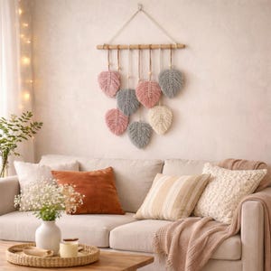 Puede incluir: Un colgante de pared de macramé con nueve diseños en forma de hoja en tonos rosa, gris y crema, suspendidos de un pasador de madera. La decoración de la pared se muestra sobre un sofá beige con cojines decorativos.