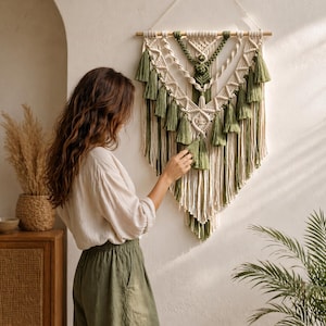 May include: A macrame wall hanging with a wooden dowel, in cream, olive green, and natural tones. The design features knotted patterns and tassels. This decorative piece adds texture and visual interest to a wall, suitable for interior decor.