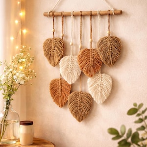 Könnte beinhalten: Makramee-Wandbehang mit acht blattförmigen Designs in Beige- und Brauntönen. Die Blätter sind an einer Holzleiste mit Naturkordel und Holzperlen aufgehängt. Ein dekoratives Stück für die Inneneinrichtung.