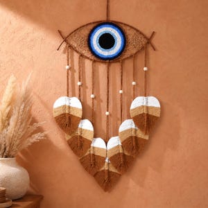 May include: A brown and white macrame wall hanging in the shape of an eye. The eye features a blue and white crocheted center, with brown string and white beads. The bottom of the hanging has six leaf-shaped macrame feathers.