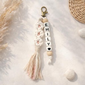 Puede incluir: Un llavero de macramé hecho a mano con el nombre "EMILY" escrito en cuentas de letras blancas. El llavero presenta borlas y detalles florales en tonos crema y rosa, sujeto a un cierre de color bronce.