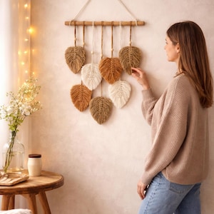 May include: A decorative wall hanging with nine woven leaf ornaments in shades of brown and cream. The leaves are suspended from a wooden dowel with a natural fibre cord. The decor is displayed against a neutral wall.