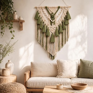 May include: A large macrame wall hanging featuring a V-shaped design with cream-colored cord and olive green accents, including multiple decorative tassels and intricate knotted patterns suspended from a horizontal wooden dowel rod.