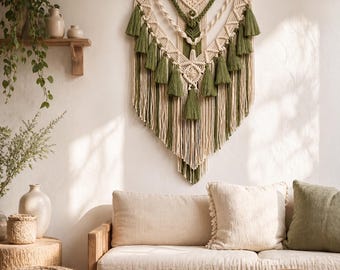 Könnte beinhalten: Ein großer Makramee-Wandbehang mit V-förmigem Design aus cremefarbenen Kordeln und olivgrünen Akzenten, bestehend aus mehreren dekorativen Quasten und komplizierten Knotenmustern, die an einer horizontalen Holzstange befestigt sind.