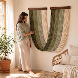 Op de afbeelding: Twee wandkleden met een houten bovenkant en verticale strengen in groen- en beigetinten. De wandkleden worden weergegeven op een lichtgekleurde muur. Het algemene thema is woondecoratie.