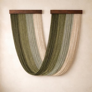 Pode incluir: Uma tapeçaria de parede com duas barras de madeira castanho-escura das quais pendem longos fios verticais. O fio cria um drapeado em forma de U, com uma transição de cor do verde-azeitona profundo para o sálvia, cinza-claro e creme.
