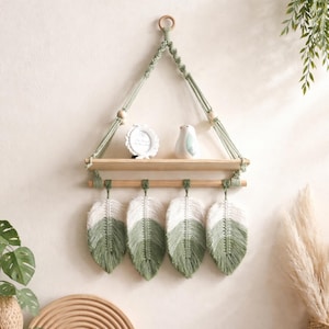 May include: A macrame wall shelf with a wooden shelf and four green and white feather decorations. The shelf is suspended by green macrame rope and features a small white picture frame and a ceramic bird figurine.