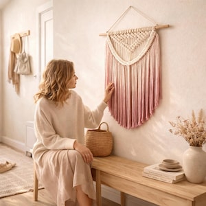 Op de afbeelding: Een macramé wandkleed met een natuurlijke houten roede en een verloop van roze en crèmekleurige katoenen draden. De wanddecoratie wordt tentoongesteld op een lichtgekleurde muur. Een vrouw raakt het wandkleed aan.