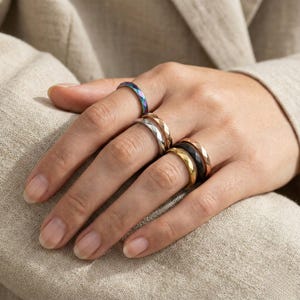 May include: A hand displays a collection of rings in various colors and finishes. The rings include iridescent, silver, gold, black, and rose gold, all with a faceted design. The rings are worn on multiple fingers, showcasing the different styles.