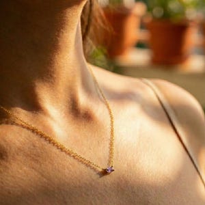 Könnte beinhalten: Eine zarte goldene Halskette mit einem kleinen, rechteckigen, lila Edelsteinanhänger. Die Kette ist fein und der Anhänger ist in einem schlichten, minimalistischen Stil gehalten. Die Halskette wird am Hals einer Person getragen, der Hintergrund ist unscharf.