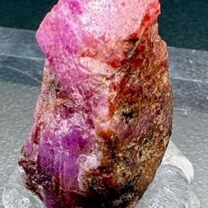 Peut inclure: Un brut de rubis de couleur rose vif, posé sur un socle transparent et texturé. La surface du rubis présente un mélange de rouge profond et de tons roses plus clairs, avec une texture brute et naturelle. Un exemple saisissant de beauté minérale naturelle.