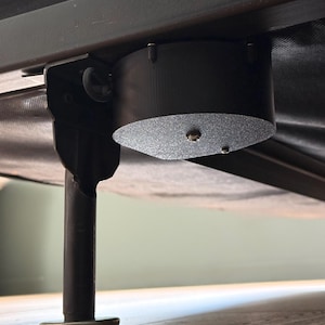May include: A close-up view of a black bed frame with a cylindrical component attached. The component has a textured, silver-colored bottom. A black metal leg with a white base is visible.