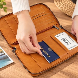 May include: A tan zipped travel organiser wallet featuring multiple interior slots for cards, a zipped pocket, and a dedicated sleeve holding a blue passport. The wallet includes a clear window slot with a card labelled RFID protected.