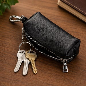 May include: A black textured leather key case with a silver-toned zipper closure and a metal swivel clasp. A silver chain is attached to the case, holding a metal ring with three keys in silver and brass tones, organized for daily use.
