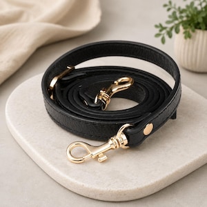May include: A black leather bag strap coiled into a circle, featuring a textured surface and polished gold-tone metal swivel lobster clasps at both ends. The strap is secured with a small gold-tone metal rivet near each of the two metal clasps.