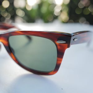May include: Tortoiseshell-patterned sunglasses with a brown and black marbled frame and dark green lenses. The frame features silver-toned metal accents at the hinges and temples, providing a classic look with a smooth, polished finish throughout.