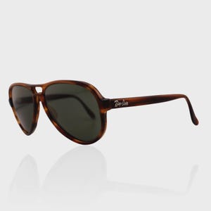 May include: Aviator sunglasses featuring a brown tortoiseshell frame and dark green lenses. The temples display the white Ray-Ban logo. These sunglasses have a classic teardrop shape with a double bridge design, providing a distinct look.