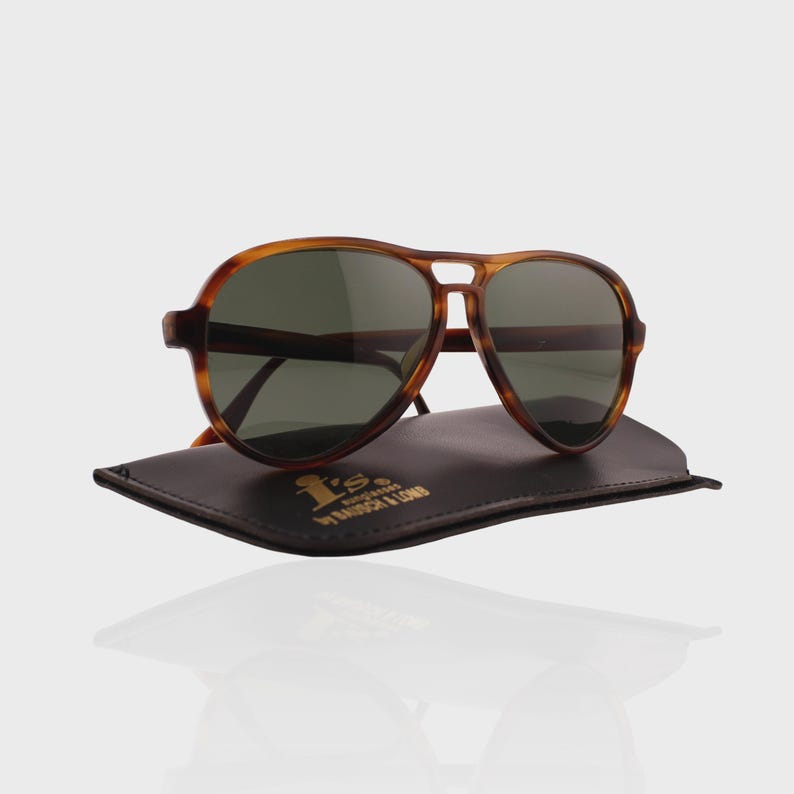May include: Aviator sunglasses with a brown tortoiseshell frame and dark green lenses. The sunglasses rest on a dark brown leather case featuring the gold text I's sunglasses by BAUSCH & LOMB. The design includes a double bridge bar.