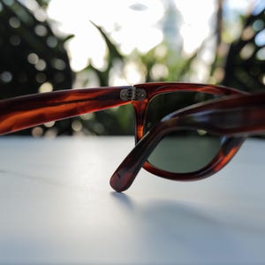 May include: Tortoiseshell-patterned sunglasses featuring a brown and amber marbled frame with dark green lenses. The frame includes a visible metal hinge mechanism connecting the temple arm to the front piece, showcasing a classic design.