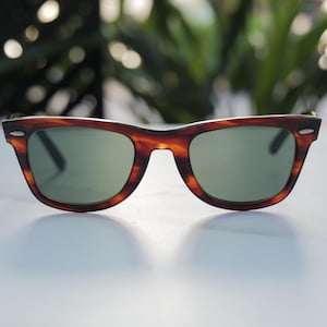 May include: Tortoiseshell-patterned sunglasses featuring a classic wayfarer-style frame with a dark brown and amber mottled finish. The lenses are a solid, muted green tint, and the frame includes small silver-toned metal accents at the outer corners.