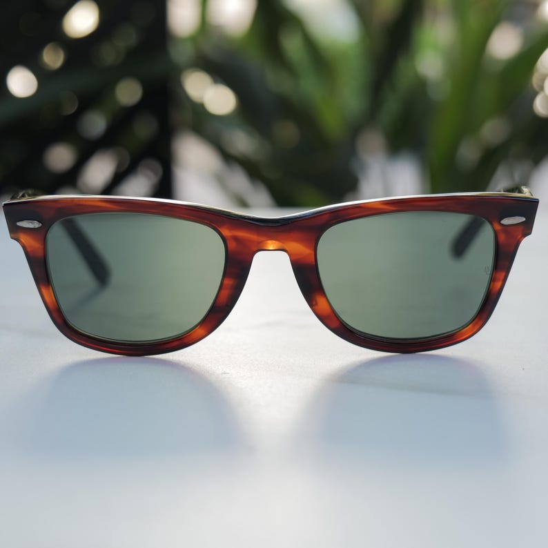 May include: Tortoiseshell-patterned sunglasses featuring a classic wayfarer-style frame with a dark brown and amber mottled finish. The lenses are a solid, muted green tint, and the frame includes small silver-toned metal accents at the outer corners.