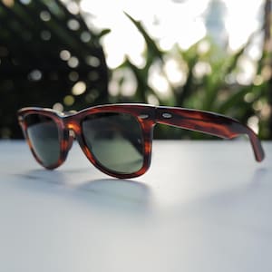 May include: Tortoiseshell-patterned sunglasses featuring a classic wayfarer-style frame with dark green lenses. The frame has silver-toned metal accents at the hinges on both sides and a small logo etched onto the right lens near the hinge.