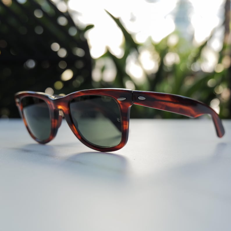 May include: Tortoiseshell-patterned sunglasses featuring a classic wayfarer-style frame with dark green lenses. The frame has silver-toned metal accents at the hinges on both sides and a small logo etched onto the right lens near the hinge.
