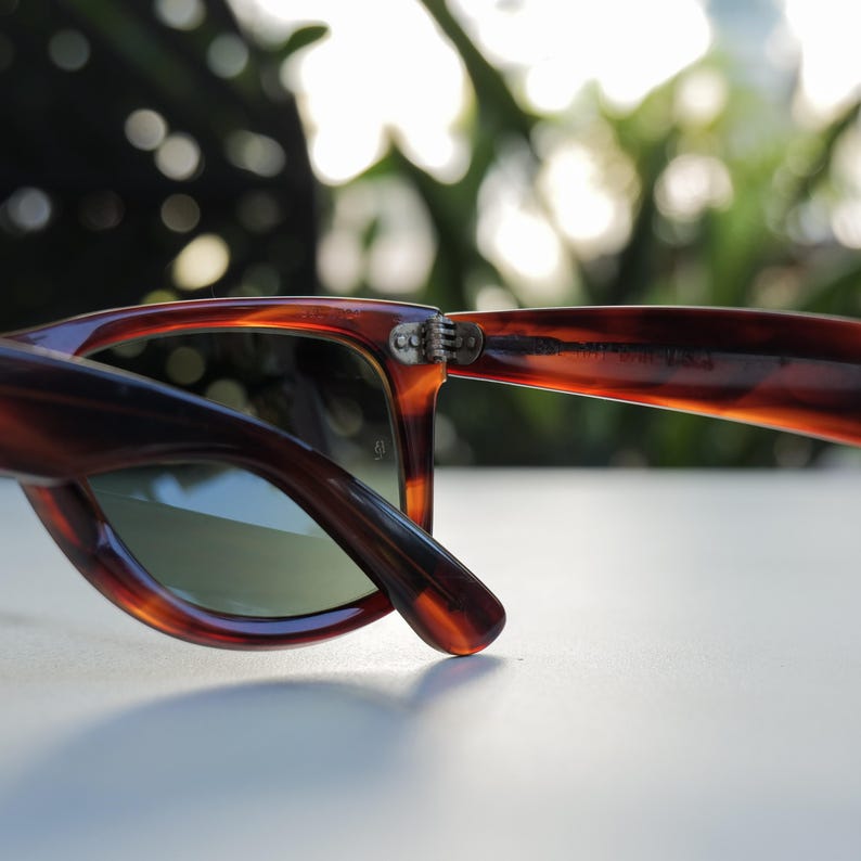May include: Tortoiseshell-patterned sunglasses featuring a brown and amber mottled frame with a glossy finish. The glasses have a metal hinge connecting the temple to the frame front, with green-tinted lenses and visible text on the inner temple.