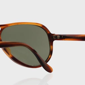 May include: Tortoiseshell-patterned sunglasses featuring a rounded frame design with dark green tinted lenses. The frame is constructed from a brown and amber mottled material, showcasing a visible metal hinge connecting the temple to the front.