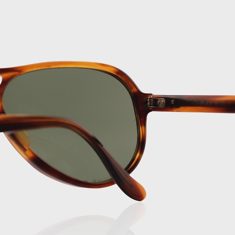 May include: Tortoiseshell-patterned sunglasses featuring a rounded frame design with dark green tinted lenses. The frame is constructed from a brown and amber mottled material, showcasing a visible metal hinge connecting the temple to the front.