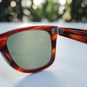 May include: Tortoiseshell-patterned sunglasses with a brown frame and green-tinted lenses. The frame features a visible metal hinge connecting the temple to the front piece, with small engraved text visible on the inner upper rim of the frame.
