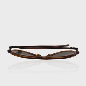 May include: A pair of folded sunglasses with a brown tortoiseshell pattern frame and dark tinted lenses. The glasses are positioned horizontally, showing the temples folded against the frame, highlighting the smooth, curved edges of the eyewear.