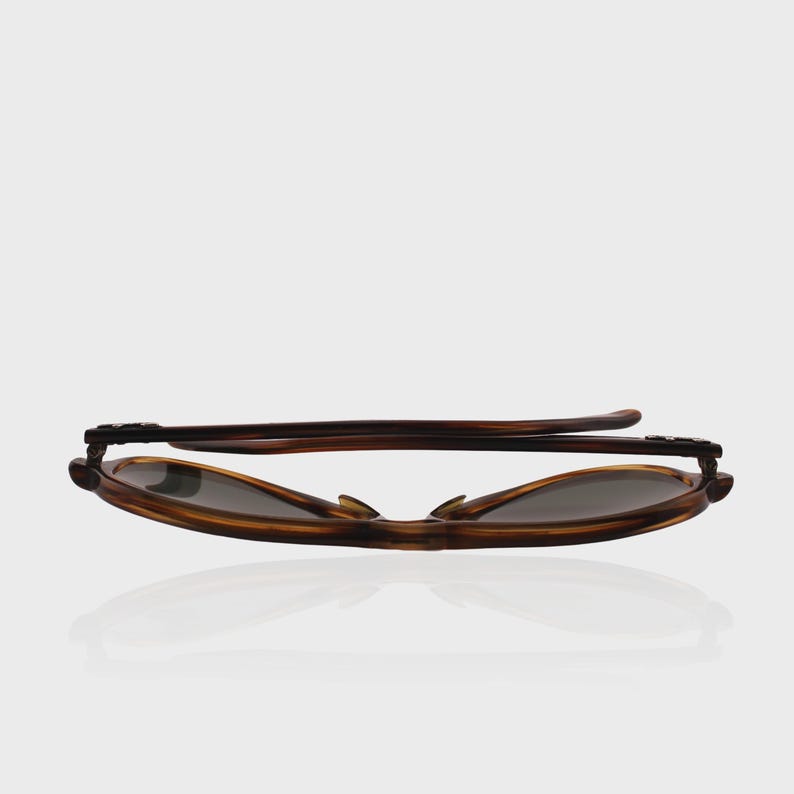 May include: A pair of folded sunglasses with a brown tortoiseshell pattern frame and dark tinted lenses. The glasses are positioned horizontally, showing the temples folded against the frame, highlighting the smooth, curved edges of the eyewear.