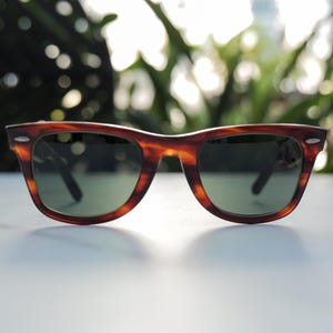 May include: Tortoiseshell sunglasses featuring a classic wayfarer frame design with dark brown and amber mottled patterns. The lenses are dark green and tinted, framed by a thick plastic rim with small silver-toned metal accents at the corners.