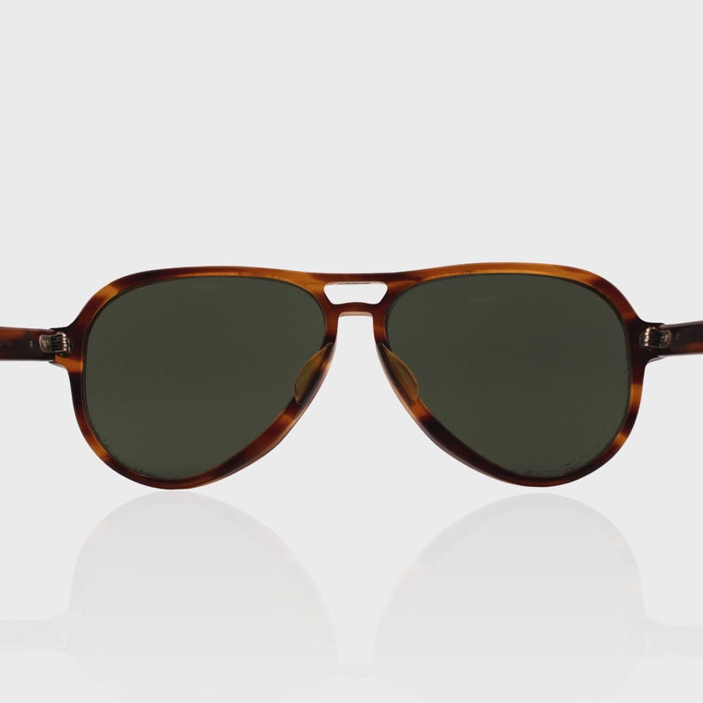 May include: Aviator sunglasses featuring a brown tortoiseshell pattern frame with a double bridge design and dark green tinted lenses. The eyewear has a classic teardrop shape with metal hinges connecting the temples to the front frame.