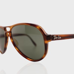 May include: Aviator sunglasses featuring a brown tortoiseshell frame and dark green lenses. The temples display a silver-toned Ray-Ban logo. The design includes a double bridge across the top of the frame, characteristic of the classic aviator style.