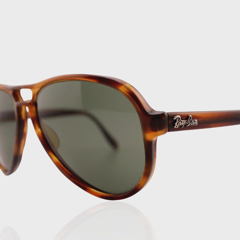 May include: Aviator sunglasses featuring a brown tortoiseshell frame and dark green lenses. The temples display a silver-toned Ray-Ban logo. The design includes a double bridge across the top of the frame, characteristic of the classic aviator style.