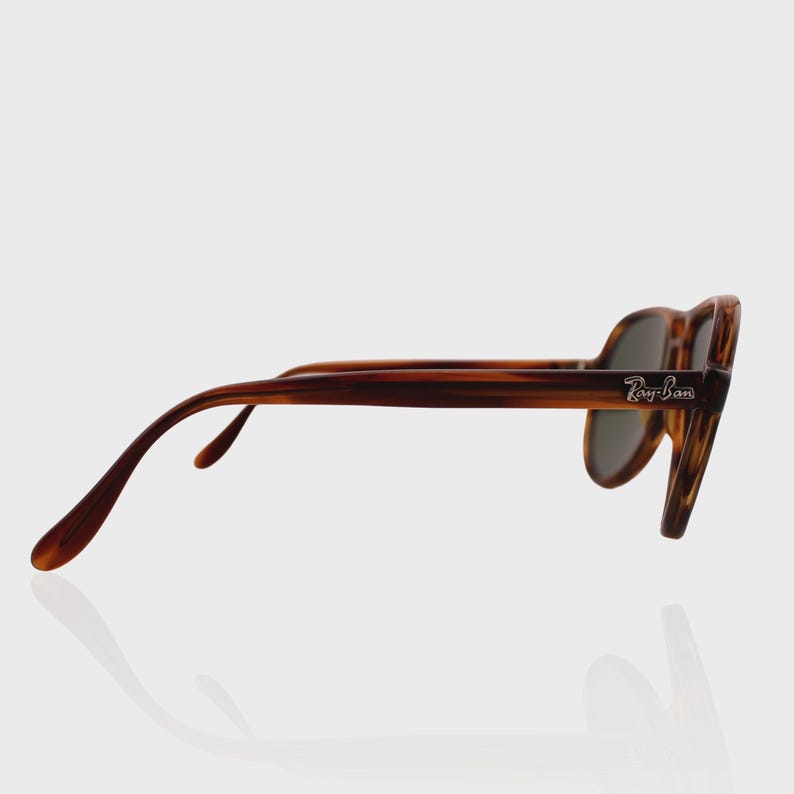 May include: Tortoiseshell sunglasses with a brown frame and dark lenses. The side of the frame features the silver Ray-Ban logo. The glasses are shown from a side profile angle, highlighting the curved temple arms and the frame's pattern.
