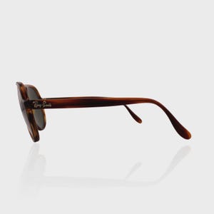 May include: Tortoiseshell-patterned sunglasses with dark lenses and a curved frame. The side of the temple features the silver-colored Ray-Ban logo. The glasses are shown from a side profile view, highlighting the brown and black mottled frame design.