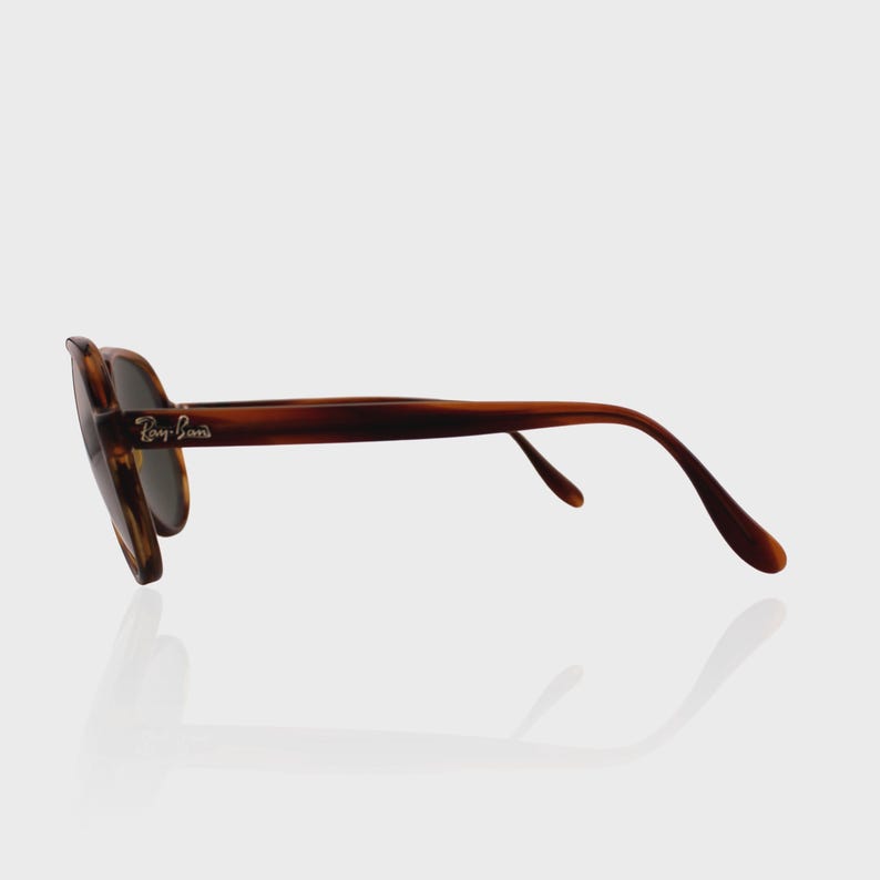May include: Tortoiseshell-patterned sunglasses with dark lenses and a curved frame. The side of the temple features the silver-colored Ray-Ban logo. The glasses are shown from a side profile view, highlighting the brown and black mottled frame design.