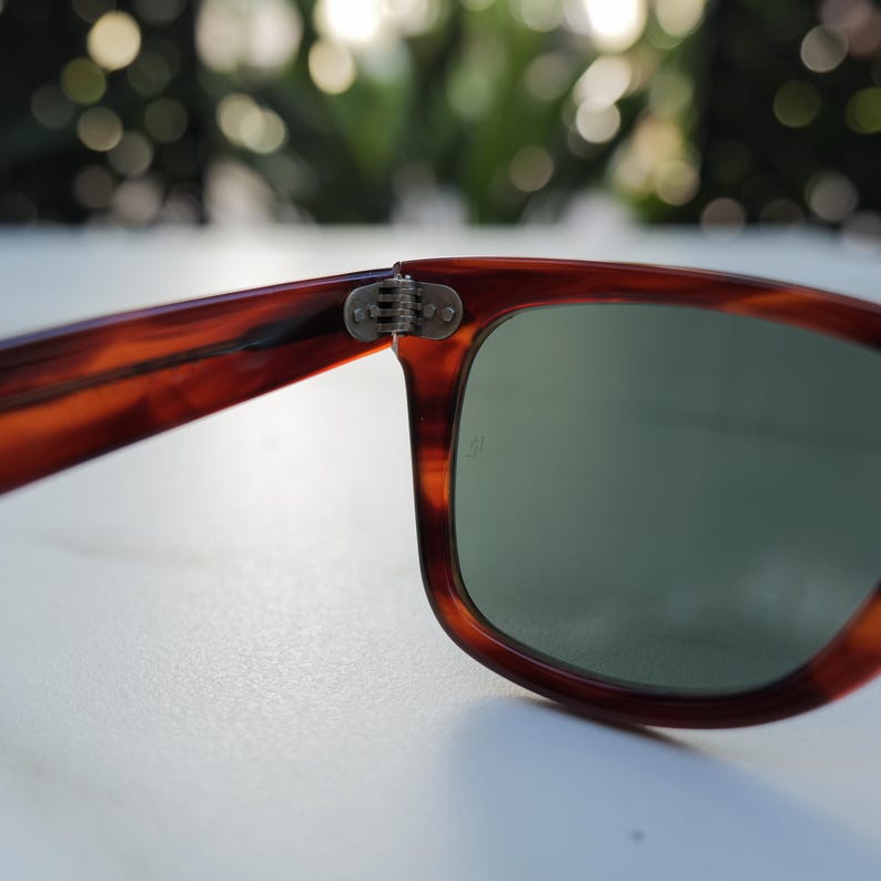 May include: Tortoiseshell-patterned sunglasses featuring a brown and amber frame with a visible metal hinge connecting the temple to the rim. The lenses are a solid, muted green color, and a small, faint logo is etched onto the right lens surface.