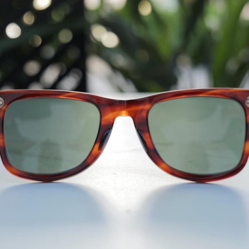May include: Tortoiseshell sunglasses featuring a classic rectangular frame design with dark brown and amber mottled patterns. The lenses are a solid, muted green tint, and the frame includes small silver-toned metal accents at the outer corners.