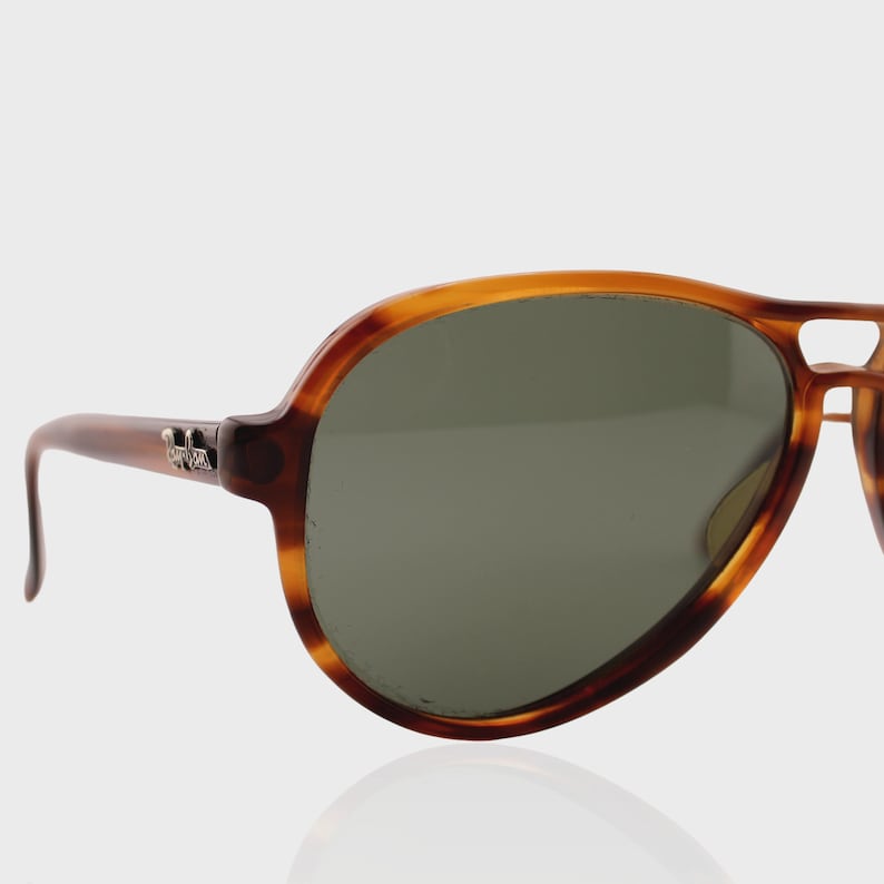 May include: Aviator sunglasses featuring a brown tortoiseshell frame with a double bridge design and dark green lenses. A silver-toned metal logo emblem is attached to the temple hinge area, providing a distinct detail to the eyewear frame.