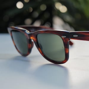 May include: Tortoiseshell-patterned sunglasses with a thick brown and black frame and dark green lenses. The frame features silver-toned metal accents at the hinges and a small logo etched into the right lens, showcasing a classic wayfarer shape.